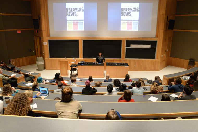 Lecture hall during a presentation. The words 'Breaking News' are visible on the screen behind the lecturer.